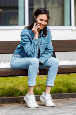 Kot ceket ve kot pantolon giyen genç bir kadın bankta oturuyor, gülümsüyor ve telefonda konuşuyor..