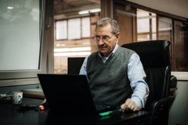 Olgun bir adam masa başında dizüstü bilgisayar kullanarak bir ofis sandalyesinde oturur..