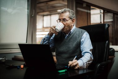 Ofisteki bir masada oturan bir adam fincandan kahve içiyor ve dizüstü bilgisayarla çalışıyor..