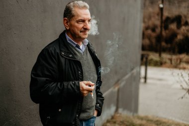 Siyah ceketli bir adam binanın yanında sigara içiyor..