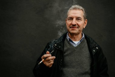 Siyah ceketli ve gri kazaklı bir adam bir sigara tutuyor ve siyah bir arka plana doğru duman üflüyor..