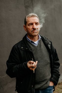 Gri saçlı ve bıyıklı yaşlı bir adam gri bir duvarın önünde durup sigara içiyor..