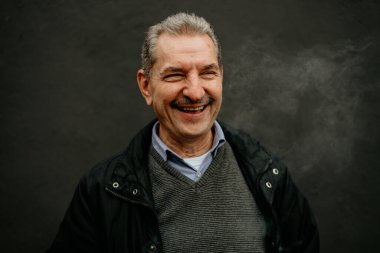 Gri saçlı ve bıyıklı bir adam gri bir kazağın üzerine siyah bir ceket giyerken geniş bir gülümseme sergiliyor..