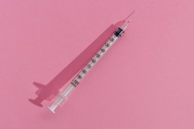 Temiz bir şırınga pembe arka planda yatıyor, gölge bırakıyor. Şırınga birim cinsinden ölçümlerle işaretlenmiş..