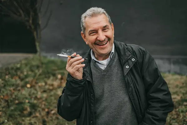 stock image A man with grey hair wearing a black jacket and a grey sweater is smoking a cigarette. He is smiling and looking at the camera.