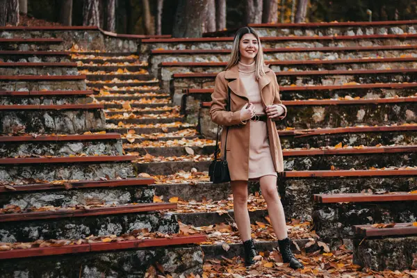 stock image A woman wearing a brown coat and a pink dress stands on a set of steps covered in fallen leaves, talking on her phone.