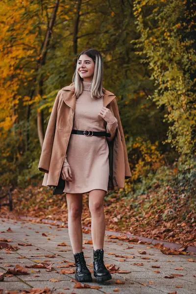 stock image A woman wearing a brown coat and a pink dress stands on a set of steps covered in fallen leaves, talking on her phone.