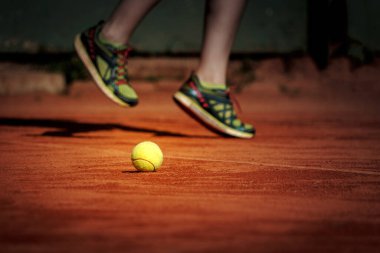 Bir tenis oyuncusu kil sahada sarı bir tenis topunun üzerinden atlıyor..