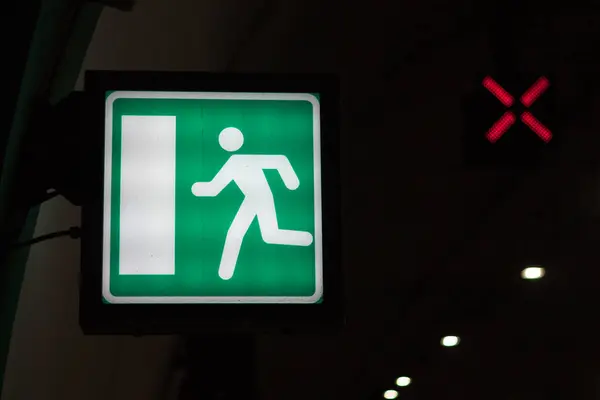 stock image A green exit sign with a white running figure is illuminated in a dark space, with a red X visible above it.