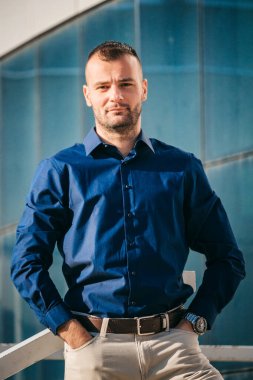 Mavi gömlekli bir adam ciddi bir ifadeyle modern cam bir binanın önünde duruyor..