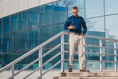 Mavi gömlekli ve haki pantolonlu bir adam modern bir binanın merdivenlerinde telefonuna bakıyor..