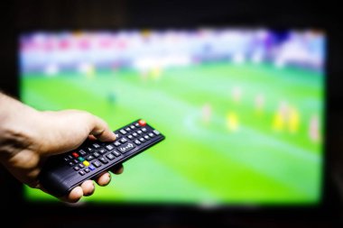 Bir kişinin eli arka planda bulanık bir futbol oyunu oynayan bir televizyon uzaktan kumandası tutuyor..