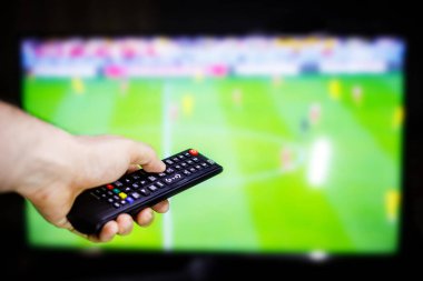 Bir el, bir futbol maçını gösteren bir televizyon ekranının önünde uzaktan kumandayı tutuyor..