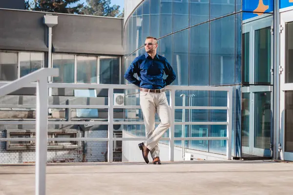 stock image A man in a blue shirt and khaki pants leans against a white railing outside a building with a modern glass facade. The sun is shining brightly, and the man is looking out at the horizon.