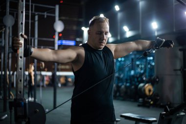 Siyah kolsuz tişörtlü bir adam geceleri spor salonunda bir kablo makinesiyle çalışıyor..