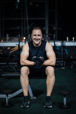 Siyah spor elbiseli bir adam spor salonundaki bir bankta oturur kolları çapraz ve siyah eldivenler giyer..
