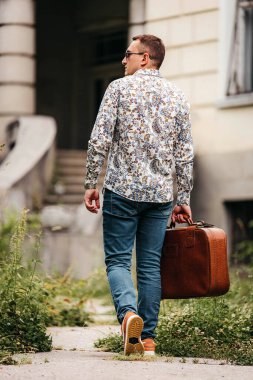 Çiçekli gömlekli ve kot pantolonlu bir adam kahverengi bir bavulla binadan çıkıyor..