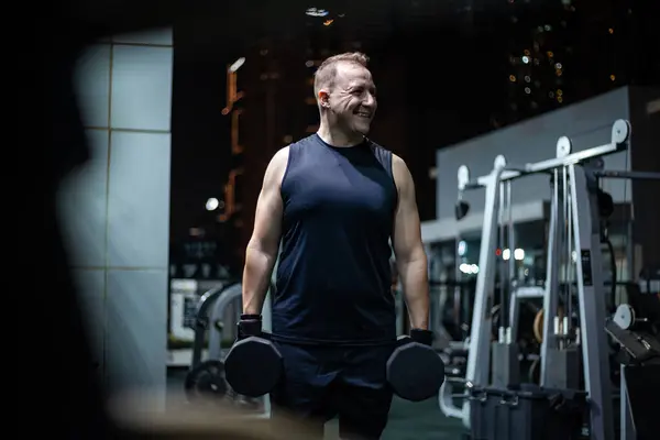 stock image A man in a black tank top lifts dumbbells while smiling in a gym at night.