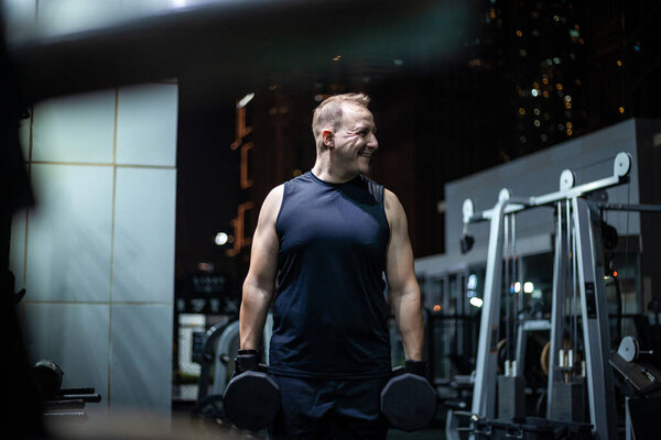 A man in a black tank top holds dumbbells in each hand, standing in a modern gym at night.