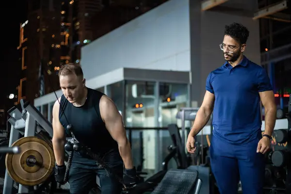 stock image A man lifts weights on a machine in a gym with his personal trainer standing by watching him.