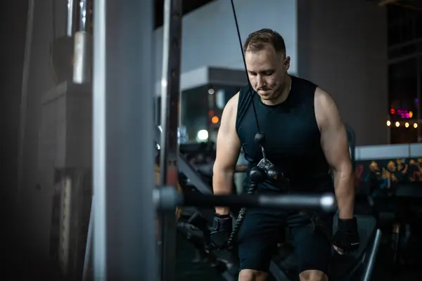 stock image A man in a black tank top and shorts uses a cable machine at a gym.