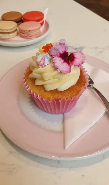 Fotoğraf, kremalı ve yenilebilir çiçeklerle süslenmiş pembe bir kağıt kaplama içinde iştah açıcı bir kek gösteriyor. Kek, yanında gümüş bir çatal bulunan pembe bir tabağa konur. Arka planda, başka bir tabakta renkli badem kurabiyeleri görüyoruz. Şey... 