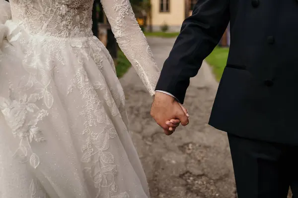 Açık havada yeni evlilerin elleri. Yürürken birbirlerine sarılan yeni evlilerin elleri. Beyaz gelinlik ve siyah takım elbise görünür..