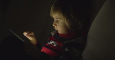 Çocuk geceleri dijital tabletin tadını çıkarıyor. Oturma odasında, kanepede oturuyor.