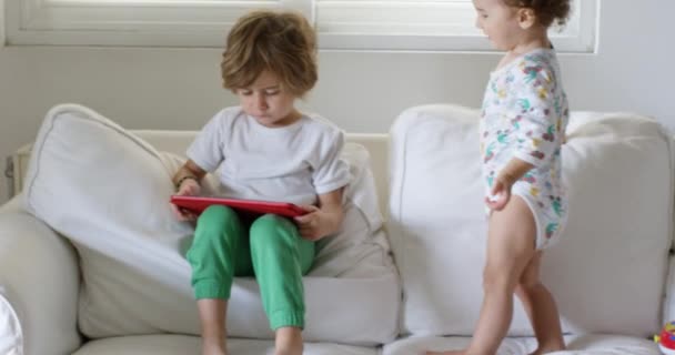 快乐的小男孩在沙发上使用平板电脑 打转社交媒体 享受呆在家里的时光 听音乐 看录像 而他一岁的弟弟在玩玩具 — 图库视频影像