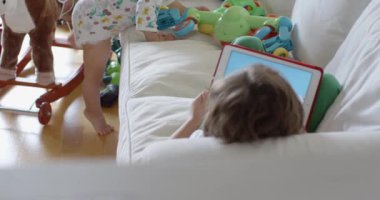 Küçük çocuk, oyuncaklarla dolu bir odada dijital tablet kullanarak beyaz bir kanepede uzanıyor..