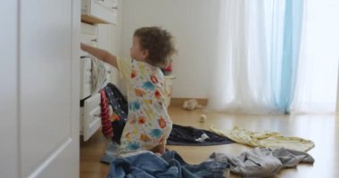 Bir yaşındaki erkek bebek dolaptan giysileri atıyor, yatak odası dağınık, içeride oynuyor ve eğleniyor, düşük açılı.