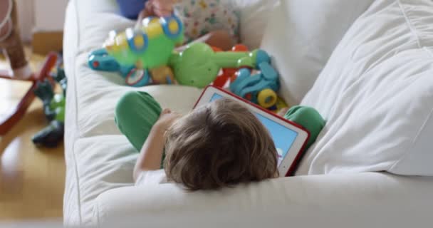 小孩子躺在白色的沙发上 用的是数码平板电脑 房间里全是玩具 — 图库视频影像
