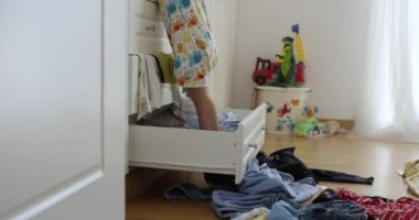 Bir yaşındaki erkek bebek dolaptan giysileri atıyor, yatak odası dağınık, içeride oynuyor ve eğleniyor, düşük açılı.