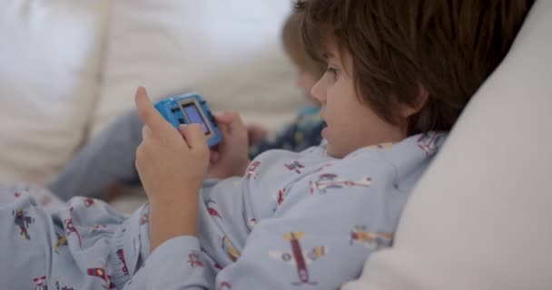 两个孩子躺在白色沙发上 专注于手持游戏装置 — 图库视频影像