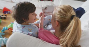 Genç anne ve çocukları oturma odasında kanepede uzanmış basılı aile fotoğraflarını izliyorlar.