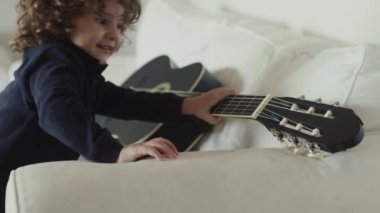 Kahverengi kıvırcık saçlı tatlı çocuk bir kanepede oturmuş gitar çalıyor..