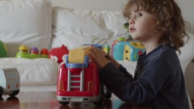 Kıvırcık saçlı bir çocuk, parlak bir odada kırmızı ve sarı oyuncak bir itfaiye arabasıyla oynuyor..