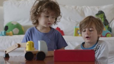 Çocuklar oyuncakları keşfediyor ve öğreniyor, birlikte eğlenceli bir aktiviteye giriyorlar, bu sırada parlak, modern bir oturma odasında dijital tablet kullanıyorlar..