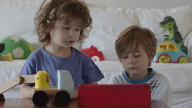 Çocuklar oyuncakları keşfediyor ve öğreniyor, birlikte eğlenceli bir aktiviteye giriyorlar, bu sırada parlak, modern bir oturma odasında dijital tablet kullanıyorlar..