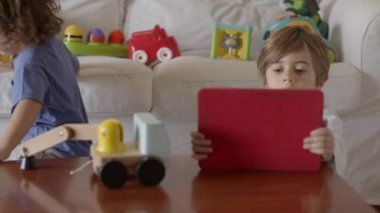 Çocuklar oyuncakları keşfediyor ve öğreniyor, birlikte eğlenceli bir aktiviteye giriyorlar, bu sırada parlak, modern bir oturma odasında dijital tablet kullanıyorlar..