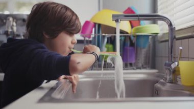 Genç bir çocuk modern bir mutfakta lavaboda suyla oynarken eğleniyor, plastik bardağı suyla dolduruyor..