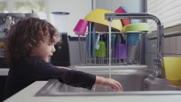 現代のキッチンでシンクで水遊びを楽しんでいる若い子供は 水でプラスチックカップを満たします — ストック動画