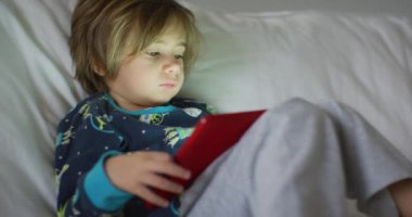 Bir çocuk dijital tablete dikkatle odaklanır, kanepeye uzanır, yüzünü aydınlatan dijital tabletin yumuşak parıltısına..