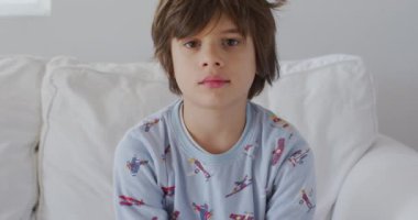 Saçları dağınık ve uykulu gözlü genç bir çocuk roket desenli pijama giyiyor ve beyaz bir koltukta oturuyor. Muhtemelen yeni uyanmış ve kameraya bakıyor..