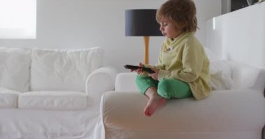 Sarı saçlı, uzaktan kumandalı tatlı bir çocuk, bir kanepenin kenarına oturmuş, televizyon programında gösterilen içeriğe odaklanmış..