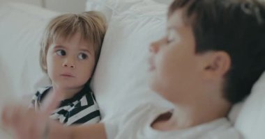 İki gencin beyaz bir kanepede oturduğu samimi bir an. Biri kardeşine tuhaf bir şekilde bakıyor ve çocukluk etkileşimlerinin özünü yakalıyor..