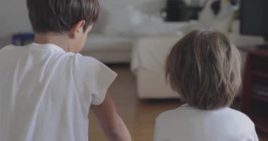 İki küçük çocuk, içeride eğleniyorlar. Gözleri dostça bakan bir yarışmaya kilitleniyorlar. Arka planda oyuncaklar var..
