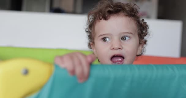 Любопытный Малыш Волнистыми Волосами Смотрит Через Край Оранжевого Манежа Смесь — стоковое видео