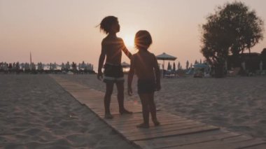 İki çocuk sahildeki sahil yolunda neşeli bir dansın tadını çıkarıyor. Batan güneşin yumuşak renklerine karşı siluetlenmiş, neşeli bir masumiyet anı yaratıyor..