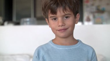 Kahverengi saçlı, mavi kazaklı bir çocuk oturuyor ve kameraya tarafsız bir ifadeyle bakıyor, gülümsüyor, mutlu görünüyor..